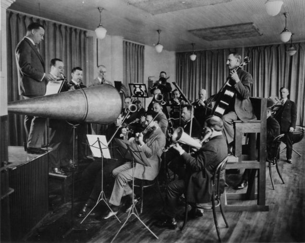Photograph of an acoustic recording session. The band is crowded around the recording horn, and most of the string players are using Stroh instruments with a horn instead of a conventional sound box.