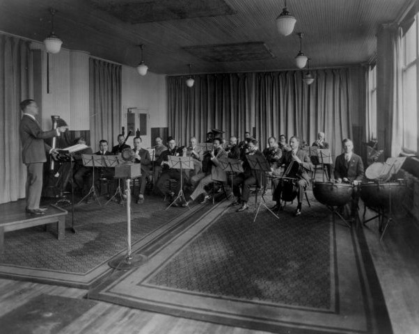 Photograph of an electrical recording session in the same studio. The band are much further back from the microphone, and are playing conventional instruments.
